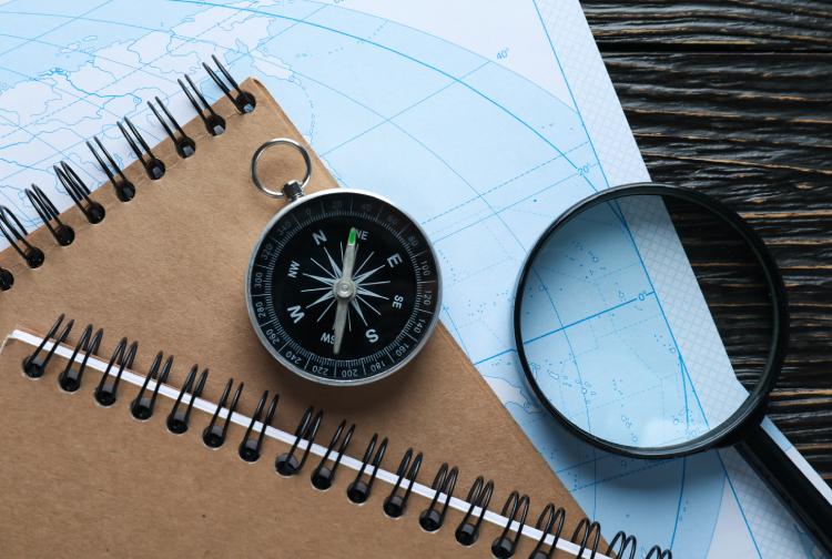 A compass, magnifying glass, and spiral notebooks placed on a world map, symbolising exploration, travel planning, or research guidance.