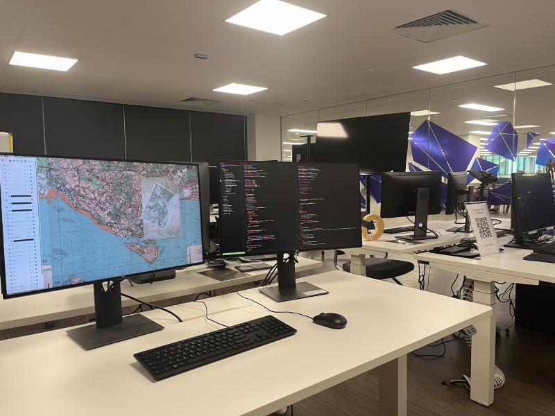 Modern classroom workspace with white desks, dual monitors displaying maps and code.
