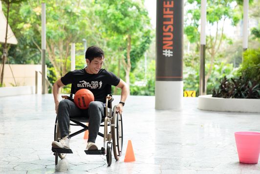 A young man in a wheel chair manoeuvring around obstacles with a basketball on his lap.